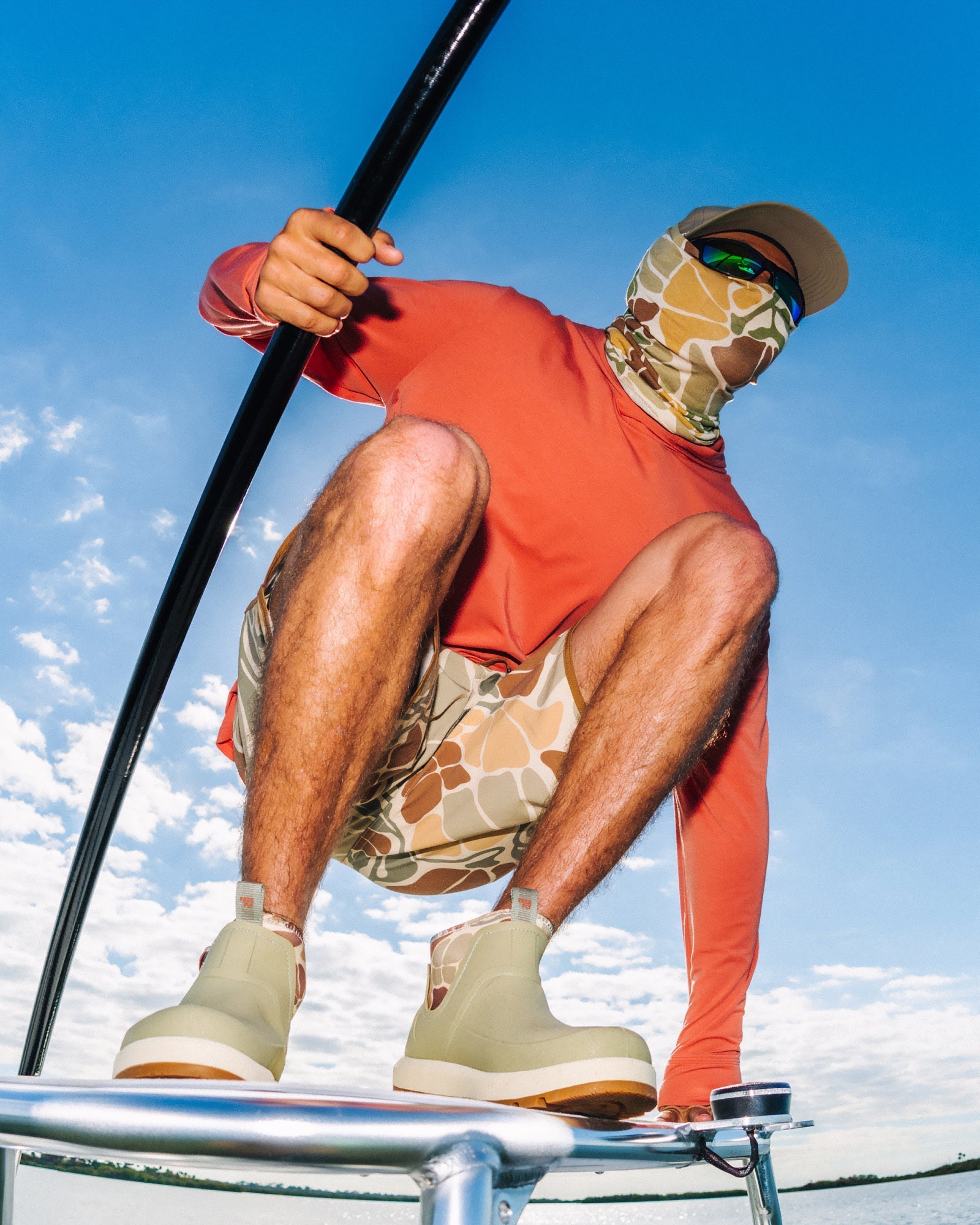 Man crouching on paddleboard wearing Bamboo Lightweight Sun Mask - Floral Camo in green and tan with orange long-sleeve top and beige water-friendly shoes facing right